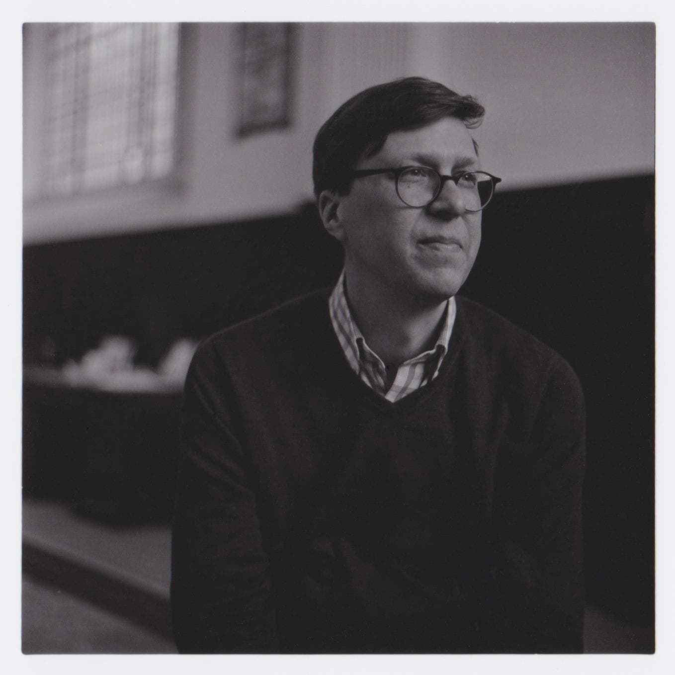 A person wearing glasses and a sweater, sitting indoors, looking off to the side in a black and white photograph.